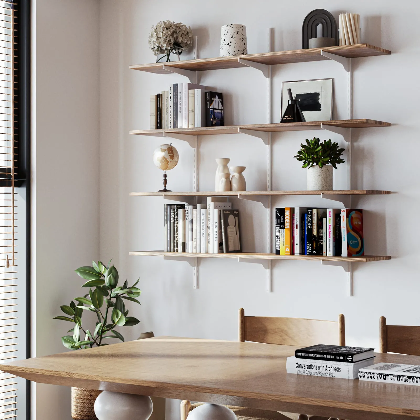 A bright home study with a pale timber wall shelving system holding books, ceramics and framed prints, a matching desk below and a chair, lit by daylight through a shuttered window.