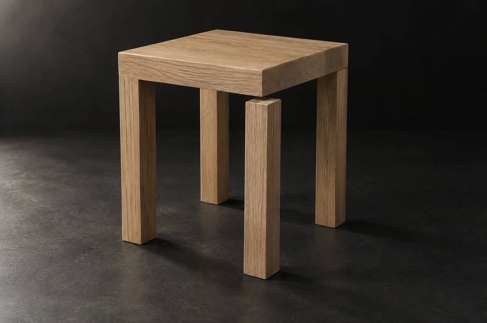 A simple oak stool on a dark studio floor, built from clean rectangular blocks. Three legs meet the seat; the fourth stands upright just off the seat's front corner, close to where it belongs but not joined to it.