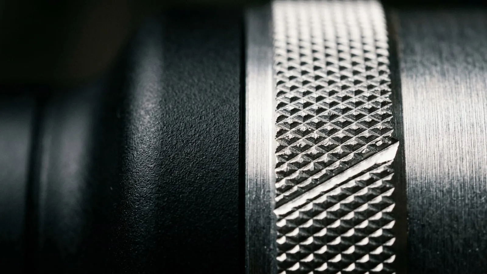 Extreme close-up of the junction where knurled brushed aluminium meets matte black polymer on a device, showing machined ridges, a crisp parting line and a chamfer catching a hard specular highlight.