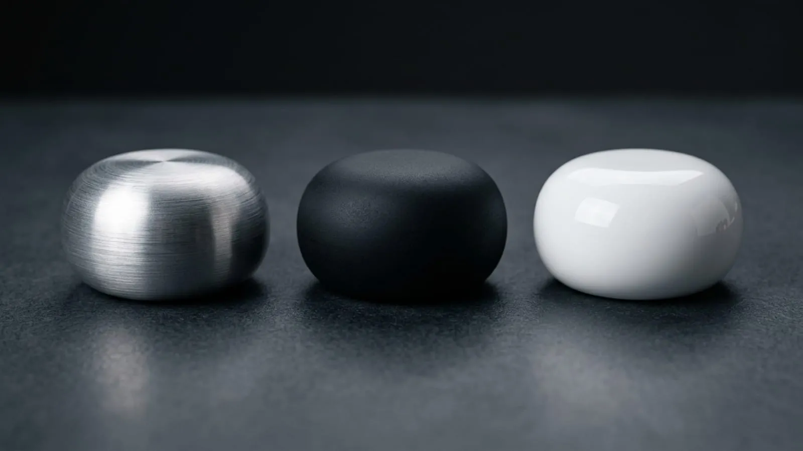 Three identical rounded forms in a row on a dark surface, the first in brushed aluminium, the second in matte black soft-touch, the third in high-gloss white.