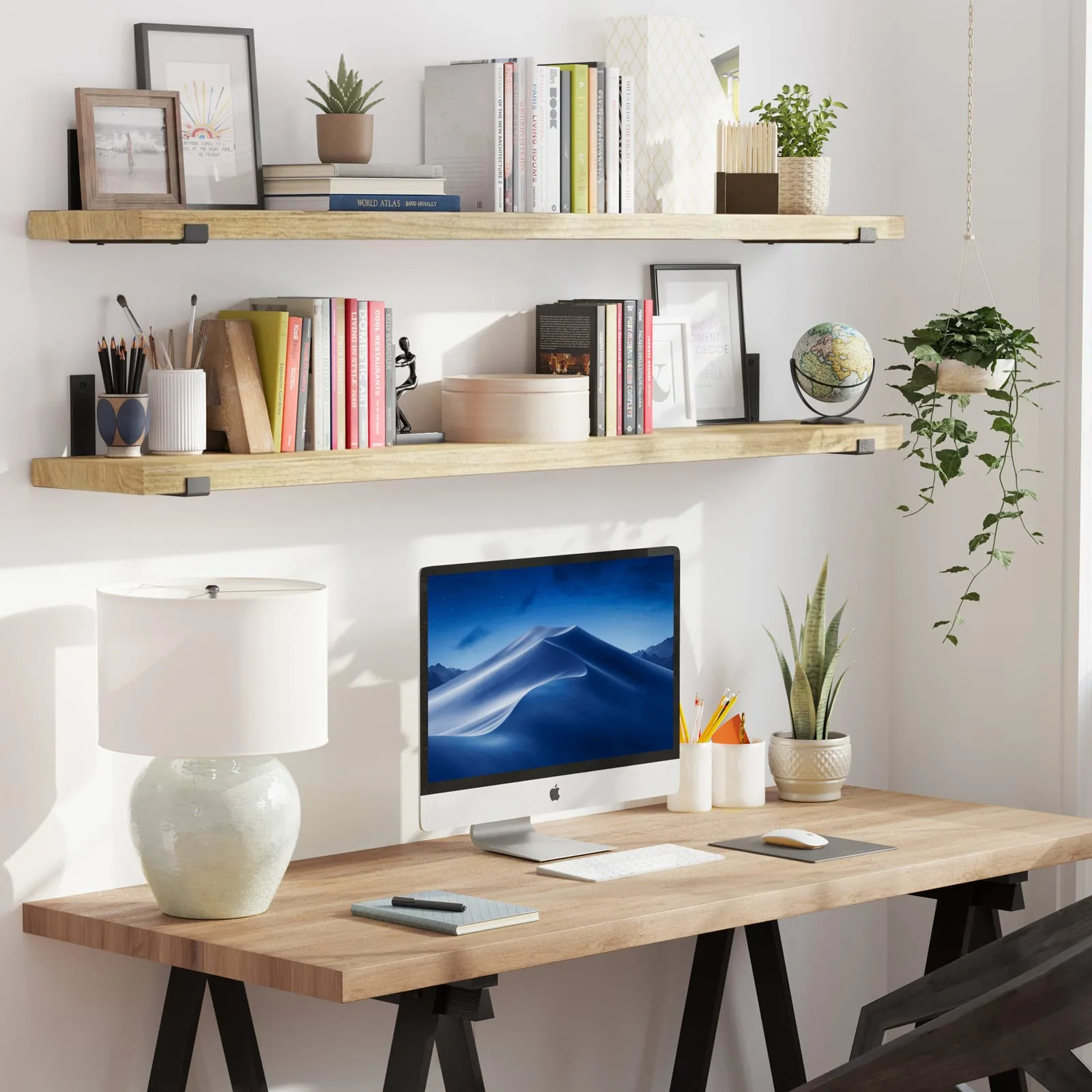 A home office corner with a light timber desk, a monitor, a ceramic lamp and two floating shelves holding books and plants, lit by soft daylight from the left.